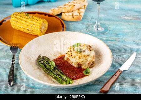 Argentinisches Entrana-Steak unter Käse, gegrilltes Rindfleisch mit Spargel, Mais und Tomatensouce auf hölzernem blauem Hintergrund Stockfoto