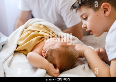 Der ältere große Bruder berührt das Neugeborene im Bett, das süße Baby unter der Decke im Bett mit glücklicher Familie, Liebe und Zusammensein im Tageslicht Stockfoto