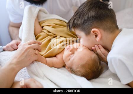 Der ältere große Bruder küsst das Neugeborene im Bett, das süße Baby unter der Decke im Bett mit glücklicher Familie, Liebe und Zusammensein im Tageslicht Stockfoto