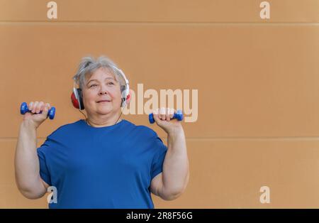 Weiße, fettleibige ältere Frau, die mit Kurzhanteln und Kopfhörern trainiert und Musik zur Gewichtsabnahme hört. Isoliert auf orangefarbenem Hintergrund Stockfoto