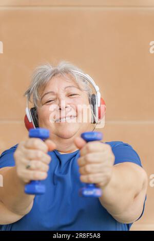 Frontalansicht einer adipösen weißhaarigen älteren Frau, die mit Kurzhanteln und Kopfhörern Musik hört. Lächelnd Stockfoto