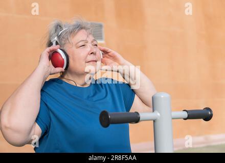 Aktive, fettleibige ältere Frau in Sportbekleidung trainiert auf einem Parkautomaten, während sie Musik über ihre Kopfhörer hört Stockfoto