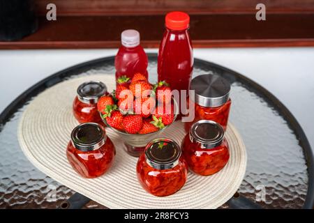 Becher mit Erdbeermarmelade, rohe Erdbeere in Schüssel, Flasche mit Sirup auf einem runden Tisch auf dem Balkon. Stockfoto