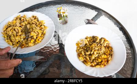 Gebackene Pasta mit Hackfleisch und geschlagenem Ei auf zwei Tellern auf einem runden Glastisch im Freien. Teil von Handbesteck. Stockfoto