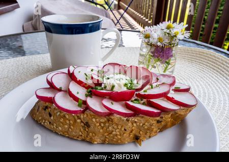 Brot garniert mit Rettich auf weißem Teller, Tasse Kaffee, Blumen auf dem Tisch, draußen. Frühstück oder Snack. Stockfoto