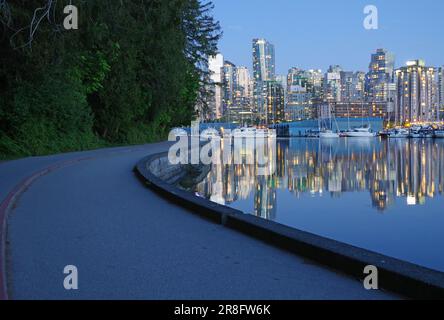 Beleuchtete Wolkenkratzer und Vergnügungsboote spiegeln sich im ruhigen Wasser wider, Teile der Sea Wall mit Radweg im Stanley Park, Dämmerung, Vancouver Stockfoto