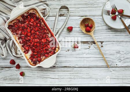 Von oben aus hat man einen Blick auf süße, hausgemachte Erdbeerkuchen oder Sonker, die in einer roten Keramikpfanne mit Schürze und Holzlöffel über einem rustikalen Tisch gebacken wurden. Stockfoto