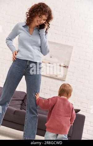 Glückliche junge Frau, die über ihr Smartphone spricht und ihr Kleinkind ansieht, die Arbeit und das Leben in Einklang bringt, moderne Elternschaft, Fernarbeit, Karriere und Familie Stockfoto