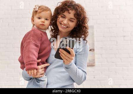 Glückliche junge Frau, die in den Armen hält, kleines Mädchen, das Smartphone benutzt, die Balance zwischen Arbeit und Leben, moderne Elternschaft, Beziehungen, Mutter Tochter bo Stockfoto