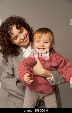 Karriere und Familie, fröhliche Frau im Anzug, die ein glückliches Kind auf grauem Hintergrund hält, beruflicher Erfolg, liebevolle Mutterschaft, Lebensstil, Multitasking, Qual Stockfoto