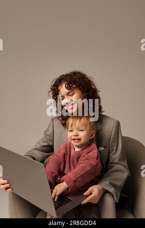 Aufbau einer erfolgreichen Karriere, ausgewogener Lebensstil, berufstätige Eltern, glückliche, lockige Frau, die mit ihrem Laptop auf einem Sessel mit Tochter, Mutter und Kind sitzt Stockfoto