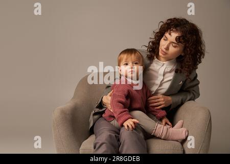 Vereinbarkeit von Beruf und Privatleben, lockige Frau im Anzug, die im Sessel sitzt, mit Kleinkindertochter, grauer Hintergrund, Multitasking, gute Familienzeit, Mo Stockfoto