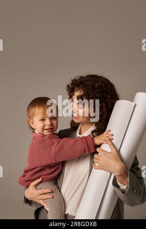 Erfolgreiche Karriere aufbauen, Multitasking-Frau, berufliche Leistungen, Zeitmanagement, fröhliche Mutter, die in Waffen hält, Tochter und Papierrollen Stockfoto