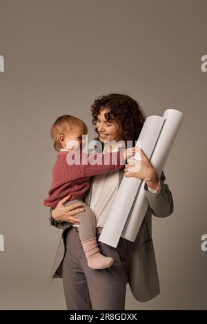 Erfolgreiche Karriere aufbauen, Multitasking-Frau, berufliche Leistungen, Zeitmanagement, glückliche Mutter, die sich in Waffen hält, Tochter und Papierrollen blu Stockfoto