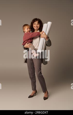 Multitasking-Frau, berufliche Leistungen, Zeitmanagement, glückliche Mutter, die sich in den Armen hält Tochter und gerollte Papierpläne auf grauem Hintergrund, Stockfoto