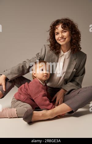 Karriere und Familie, ausgewogener Lebensstil, fröhliche Mutter mit lockigem Haar, die kleine Tochter auf grauem Hintergrund umarmt, gute Zeit, moderne Erziehung, Bu Stockfoto