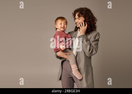 Moderne Erziehung, erfolgreiche Karriere, befähigte Frau, fröhliche Mutter, die auf dem Smartphone spricht und die kleine Tochter auf grauem Rücken in Armen hält Stockfoto