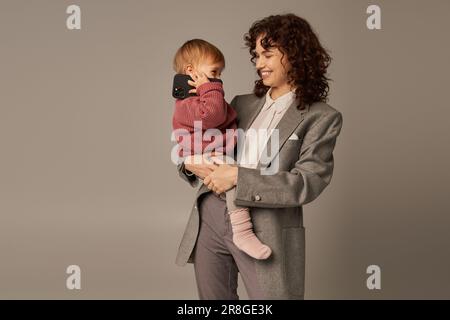 Moderne Erziehung, erfolgreiche Karriere, Gleichgewicht zwischen Arbeit und Leben, fröhliche Mutter, die Kleinkinder mit Smartphone auf Gre in den Armen hält Stockfoto