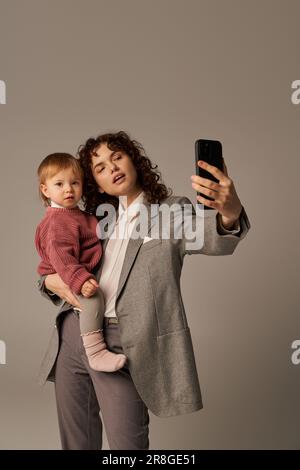 Moderne Erziehung, erfolgreiche Karriere, befähigte Frau, lockige Mutter, die Selfie auf dem Smartphone macht und Kleinkindertochter in den Armen hält, auf grau b Stockfoto