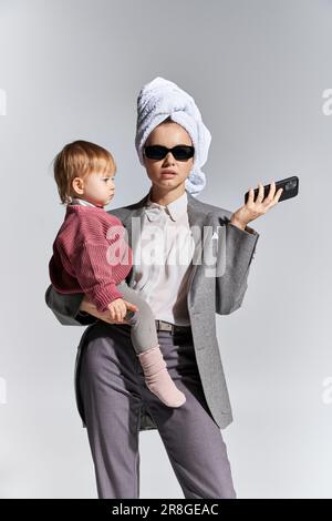 Karriere und Familie, Multitasking, Frau in Sonnenbrille, die in den Armen hält Kleinkindertochter und mit Handtuch am Kopf stehend, Geschäftsfrau in formeller Kleidung Stockfoto