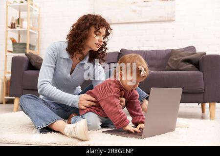 Moderne berufstätige Eltern, engagiert sich für das Kind, Gleichgewicht zwischen Arbeit und Leben, lockige Frau, die ein Mädchen in der Nähe eines Laptops unterstützt, moderne Elternschaft, Multitasking-woma Stockfoto