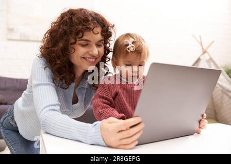 Multitasking-Frau, Freiberufler, moderne berufstätige Eltern, die Arbeit und das Leben in Einklang bringen, glückliche Frau, die ein Notebook im gemütlichen Wohnzimmer benutzt, moderne Elternschaft, eingebaut Stockfoto