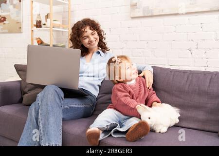 Multitasking-Frau, freiberuflich, lockige Frau, die mit einem Laptop und einem kleinen Mädchen auf dem Sofa in einem gemütlichen Wohnzimmer sitzt, moderne Elternschaft, erfolgreiches Bauen Stockfoto