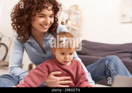 Multitasking-Frau, engagiert sich für Kind, Arbeit und Leben, lockige Frau, die das kleine Mädchen in einem gemütlichen Wohnzimmer unterstützt, moderne Eltern, liebevoll Stockfoto