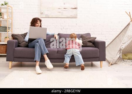 Multitasking-Frau, Freiberuflerin, lockige Frau, die ihren Laptop benutzt und auf der Couch neben der Kleinkindertochter sitzt, in einem gemütlichen Wohnzimmer, moderne Eltern, Gebäude Suc Stockfoto