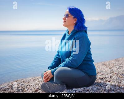 Eine Frau trägt einen hellblauen Hoodie und Jeans in Übergröße. Blauhaarige Frau draußen im Park am Strand von Antalya. Kleidungsmodell, leerer Platz für Text o Design. Stockfoto