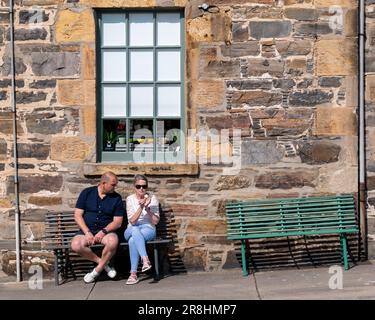 18. Juni 2023 Cullen, Moray, Schottland. Das ist ein Mann und eine Frau, die auf einer Straßenbank sitzen. Die Frau isst ein Eis, während der Mann es sich anschaut. Stockfoto