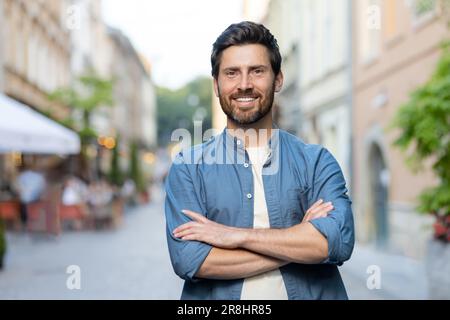 Porträt eines erwachsenen, erfolgreichen Mannes in der Stadt, der Kerl lächelt und mit gekreuzten Armen in die Kamera schaut, auf einer Reise durch die Abendstadt. Stockfoto