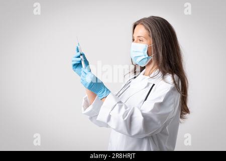 Professionelle leitende Ärztin im Mantel, Schutzmaske, Handschuhe mit Spritze und Impfstoff posieren auf hellem Hintergrund Stockfoto