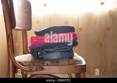 Ein Stapel gefalteter, sauberer Kleidung liegt auf einem Stuhl im Schlafzimmer in der Nähe des Fensters, Kleidung Stockfoto