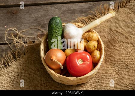 Kartoffeln Zwiebeln Tomaten Gurke Knoblauch Ernte aus dem Garten liegt in einem Korb aus Korb auf einem Holztisch, Gemüse im Hintergrund, Gemüsebacke Stockfoto
