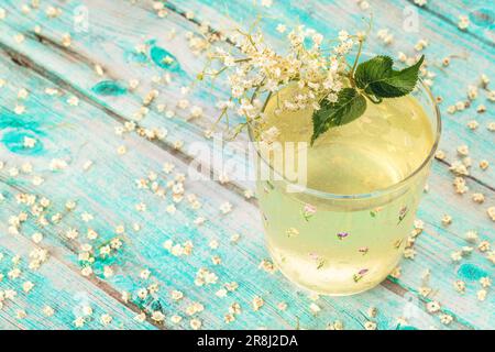 Zwerg-Holunderbeersaft, Holunderbeersirup in einem Glasbecher mit Blüten und Haufen Holunderbeeren und Holunderblättern, frischer Sommercocktail. Stockfoto