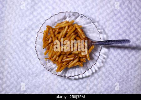 Aus einem großen Blickwinkel können Sie leckere Pasta in einer Glasschüssel betrachten, wobei der Löffel auf einem strukturierten weißen Hintergrund isoliert ist. Negativer Bereich zum Schreiben von Text Stockfoto