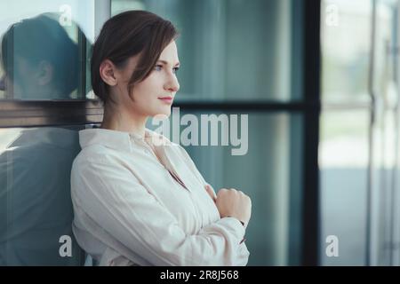 Eine selbstbewusste, wunderschöne junge weiße Frau steht am Bürogebäude, lehnt sich an eine Glaswand und denkt bewusst zur Seite Stockfoto