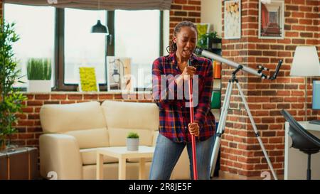 afroamerikanische Hausfrau, die Musik hört und den Boden der Wohnung mit Wischmopp und Waschlösung wischte. Junge, glückliche Frau, die tanzt und singt, Spaß beim Frühjahrsputz hat. Stockfoto