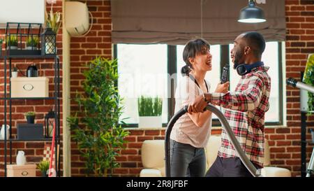 Ein vielseitiges Paar, das das Wohnzimmer mit Staubsauger und Mopp putzt, singt und Musik hört. Junge, glückliche Partner, die Frühjahrsputz genießen, Apartmentböden saugen. Stockfoto