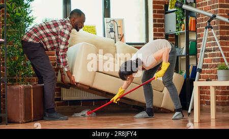 Ein vielseitiges Paar, das Holzfliesen im Wohnzimmer wäscht, ein Freund, der der Freundin hilft, den Boden unter der Couch zu reinigen. Junge Hausfrau wischte den Wohnungsboden mit Reinigungsmittel. Handgeführte Aufnahme. Stockfoto