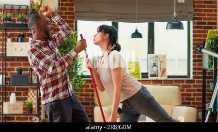 Ein modernes lächelndes Paar, das tanzt und putzt, Wisch- und Staubsauger im Wohnzimmer, Holzböden. Junge positive Partner, die Spaß mit Tanzbewegungen haben, die Hausarbeit genießen. Handgeführte Aufnahme. Stockfoto