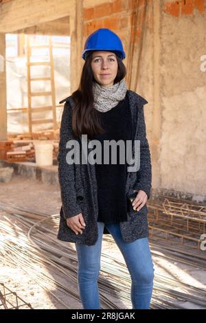 Vertikales, durchgehendes Porträt einer Architektin, die auf einer Baustelle in die Kamera schaut und einen blauen Schutzhelm trägt. Stockfoto