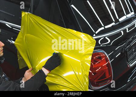 Spezialist für Fahrzeugverpackungen, der das schwarze Auto des Kunden mit neongelber oder grüner Vinylfolie beschichtet. Großer Blickwinkel. Indoor-Aufnahme. Hochwertiges Foto Stockfoto