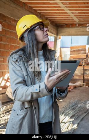 Vertikales Porträt einer Architektin mit gelbem Schutzhelm, die mit ihrem Tablet in der Hand auf die Baustelle schaut. Inspektorin mit Tisch Stockfoto
