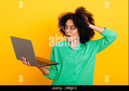 Freudig überraschte brasilianische oder afroamerikanische lockige Frau, in einem grünen Hemd, mit offenem Laptop in der Hand, sieht verblüfft darauf, hält Hand an Kopf, steht auf isoliertem gelbem Hintergrund Stockfoto