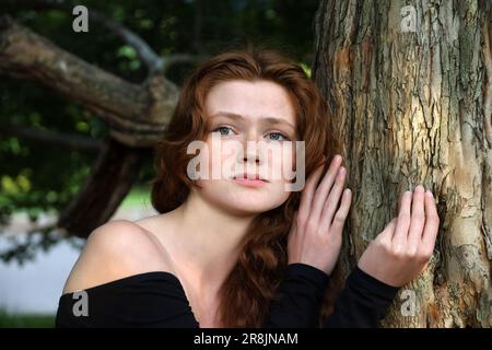 Ein Mädchen mit langen roten Haaren und Sommersprossen steht neben dem Baum im Sommerpark. Depression und traurige Stimmung Stockfoto