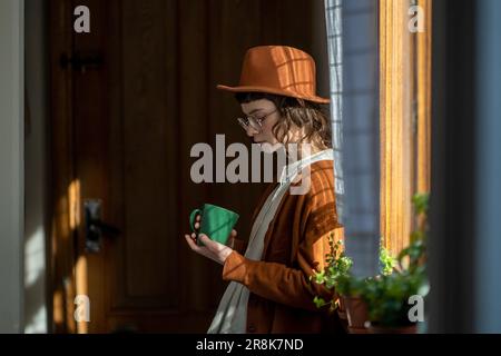 Trauriges, deprimiertes Gen Z-Mädchen, das neben dem Fenster steht und eine Tasse Tee hält, sich melancholisch anfühlt Stockfoto