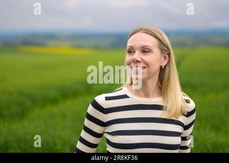 Blonde Frau mittleren Alters, die vor einem grünen Feld steht und nebenbei freundlich aussieht Stockfoto