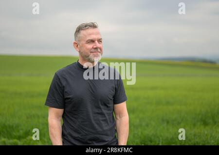 Ein grauhaariger Mann im mittleren Alter, der ein schwarzes Hemd trägt, steht vor einem Feld und sieht seitlich freundlich aus Stockfoto